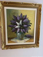 Full view of framed floral oil painting on canvas showing purple and white flowers with green leaves in a vase on a table with tablecloth, ornate gold frame.
