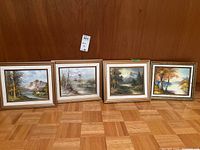 Four framed landscape paintings displayed on floor against wooden panel wall, showing full set and framing details