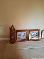 Two framed signed paintings with urban scenes and two gold painted flower plaques leaning against the wall on the floor.