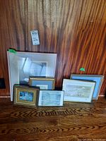 Five framed prints leaning against a wood-paneled wall on hardwood floor showing various sizes and frame styles