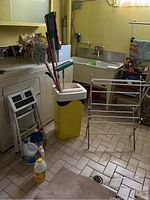 Photo showing yellow garbage bin with mop bucket attached, assorted mops inside, small step stool, and bottles of cleaning supplies on tiled floor near washing machine and laundry sink.