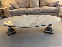 Front perspective view showing oval white veined stone top on twin ornate pedestal bases with sofa background