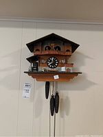 Full view of the wooden chalet cuckoo clock mounted on wall with three pinecone weights hanging and detailed base including water wheel and stair motif.