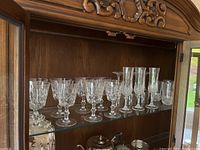 Wide view of crystal stemware including wine glasses, champagne flutes and highball glasses arranged inside wooden cabinet.