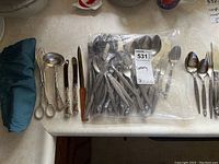Photo showing assorted stainless steel cutlery including spoons, forks, and knives with decorative handles, plus ladle and tongs on a countertop.