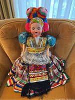 Full front view of the doll sitting on a chair showing colorful folk costume, lace apron, and pom-pom headpiece.
