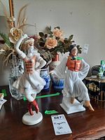 Two ceramic folkloric figurines on a table with decorative vases and flowers in background.