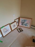 Six framed floral art pieces leaning against a wall on carpet. Varied sizes and shapes including rectangular and oval frames, with visible price stickers.