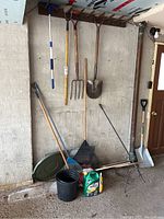 Overview of multiple garden tools including forks, shovels, broom, containers, and garden care product in used condition, arranged in garage.
