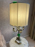 Single lamp close-up showing 5 bulb holders under cream scalloped lampshade with pink fabric tape marker on top, metal and green glass base on white marble top.