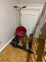 Photo shows Toro Lawn Baron seeder with large red hopper, T-shaped steel handle, two translucent hanging bird feeders with different colored tops, and a black Toro handheld leaf blower all placed on parquet floor.