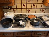 Countertop with multiple pots and fry pans, including black and copper-colored pans, a white salad spinner, placed on a stove surface under kitchen cabinets with floral tile backsplash.