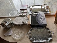 Overview of serving ware including metal trays, crystal bowl, glass plates, coasters, covered cake plate, and tablecloth
