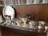 Full view of silver plate tea set on rectangular tray, red glass candle holders, silver plate trays, salt and pepper shakers, and glasses on glass shelf