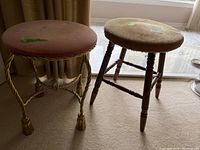 Photo showing two stools side by side: one has a twisted metal base with tassel feet and a round brown seat cushion; the other has a wooden turned leg base with a light beige cushioned seat showing stains.