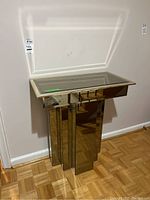 Full view of the glass table against the wall with parquet flooring, showing the vertical reflective metal panel base.