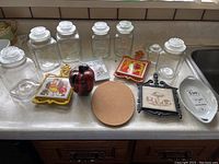 Seven glass canisters, ceramic apple bank, four ceramic trivets, and one cork trivet on kitchen counter