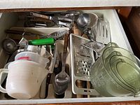 Full top view of kitchen utensil drawer showing stacked nested measuring cups, metal graters, knives with dark handles, ladle, rolling pin, plastic spatulas and spoons.