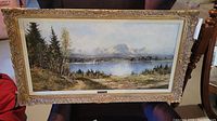 Full view of oil painting framed in ornate gold frame showing lake with sailboats and surrounding natural scenery.