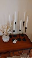 Six black metal candle holders with white taper candles arranged on wooden surface next to ceramic vase.