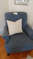 Front left angle of Clayton Marcus blue oversized armchair with beige pillow, showing armrest and wooden caster leg.