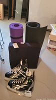Photo showing two rolled yoga mats (purple and dark gray), black back support cushion with handles, and men's size 10 ice skates.