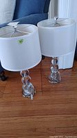 Two matching table lamps placed side by side on hardwood floor near blue upholstered chairs, showing entire lamps including white shades and clear glass orb bases.