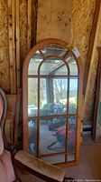 Front view of a tall, arched wood framed mirror with grid design in the glass, showing the entire mirror leaning against a wooden wall.