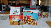Wide view of all boxed kitchen appliances and tools on table: Imperia pasta maker, Cuisipro yogurt/cheese maker, Thomson and Taylor Velox tomato press, Browne rotary cheese grater.