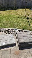Two stacks of gray cement pavers, each stack containing three pavers, placed on a paved patio surface next to a garden bed with decorative rocks. Green grass visible in background.