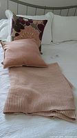 Wide image showing two decorative pillows and a folded pink knitted throw blanket laid on a bed with a white bedspread, illustrating the lot contents.