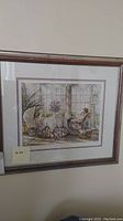 Framed print in wood frame showing woman seated by table near window with floral and vintage decor.