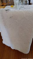 Photo showing the folded white quilt with lace trim at the edges, hanging over a surface with some glassware on top.
