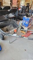 Seed spreader in a garage with black hopper, orange dial control, digital labels, metal frame, handle, and wheels visible.