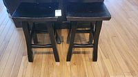 Photo showing two black wooden stools side by side on a wood floor with rectangular seats and cross-bracing support bars.