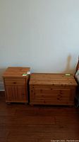 Photo showing both the wooden table with drawer and cabinet door alongside the wooden storage chest with a hinged lid, both placed against a white wall on wooden flooring.