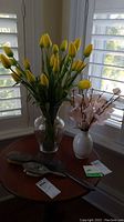 Wide view showing all items on round wooden table by window. Includes glass vase with yellow tulips, white ceramic vase with pink flowers, two vintage brushes and one hand mirror.