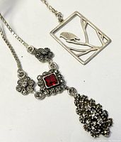 Close-up of two silver necklaces, one with an open bird and branch design pendant, the other with a detailed floral silver pendant with a red stone.