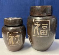 Two dark brown pottery urns with lids and etched Korean characters on the front, shown side by side on a white surface.