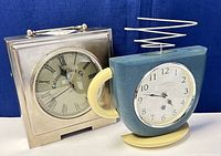 Two vintage mantle clocks side by side on a dark blue background including a silver-tone Colonial Clock Co. clock and a blue and cream novelty Ingraham clock with wire antenna.