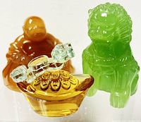 Three small carved resin figurines: amber Buddha, green foo dog, gold ingot with clear accents, arranged side by side on white background.