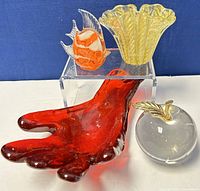 Four assorted vintage hand blown glass pieces displayed together: a red glass hand, a yellow ruffled vase, a orange and white striped fish, and a clear apple with gold leaf.