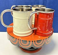 Four vintage tin beer mugs (3 cream, 1 red) arranged on top of the footed ceramic cake plate, showing mugs' shape, handles, and interior stainless steel lining.