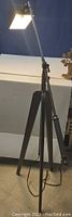 Full view of the modernist tripod floor lamp with the light on, showing the lamp head, adjustable arm, and tripod legs.