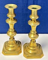 Pair of old brass candleholders standing side by side against blue background showing overall shape and hexagonal base.