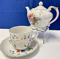 Photo showing large ceramic teacup with 'FATHER' inscription and ceramic saucer with colorful berry design in front; vintage ceramic musical teapot with pink and multicolor floral pattern behind on a clear stand.