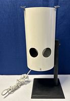 Vintage Hubbell table lamp with a cylindrical white metal shade and two circular cutouts, mounted on a black metal rod and square base. White cord attached.