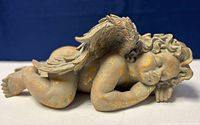 Side view of a vintage sleeping cherub sculpture showing detailed curled hair and feathered wings with a distressed light brown finish and minor flea chips.