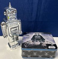 Photo showing the ceramic robot coin bank next to the Star Wars tin lunchbox with Darth Vader printed on the lid and Star Wars logos on the sides.