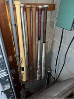 Photo showing eight baseball bats of wood and metal in worn condition, standing upright in corner.